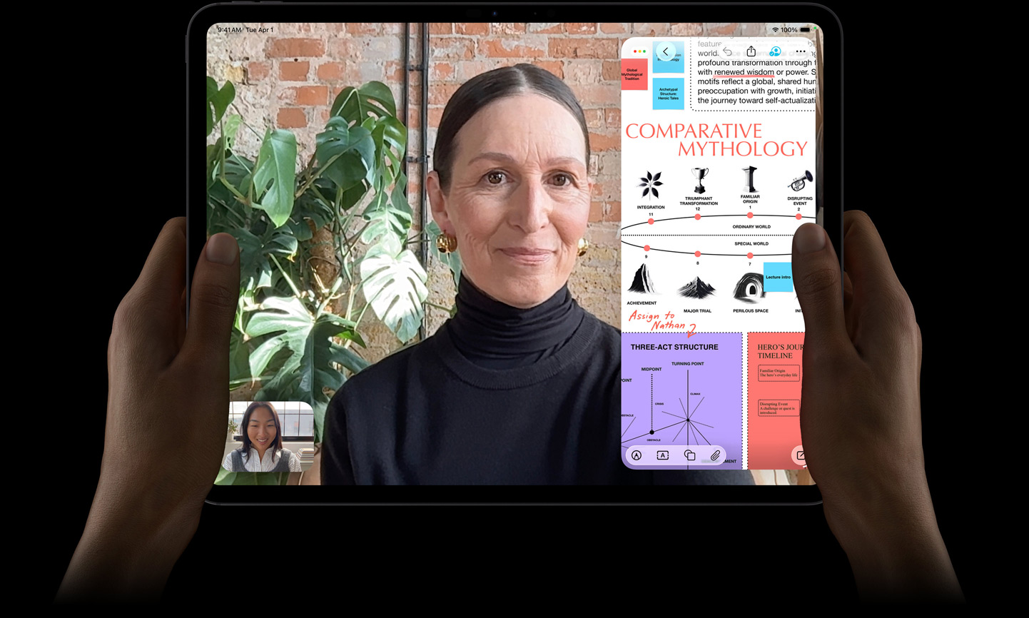 En iPad Pro i fargen stellarsvart vises forfra. To hender holder den i liggende retning. Enheten har avrundede hjørner og svart kant rundt skjermen, som viser en direkteoversatt FaceTime-samtale og en utdanningsapp. Utdanningsappen viser en presentasjon om «Comparative Mythology» med tekst, diagrammer og håndskrevne notater.