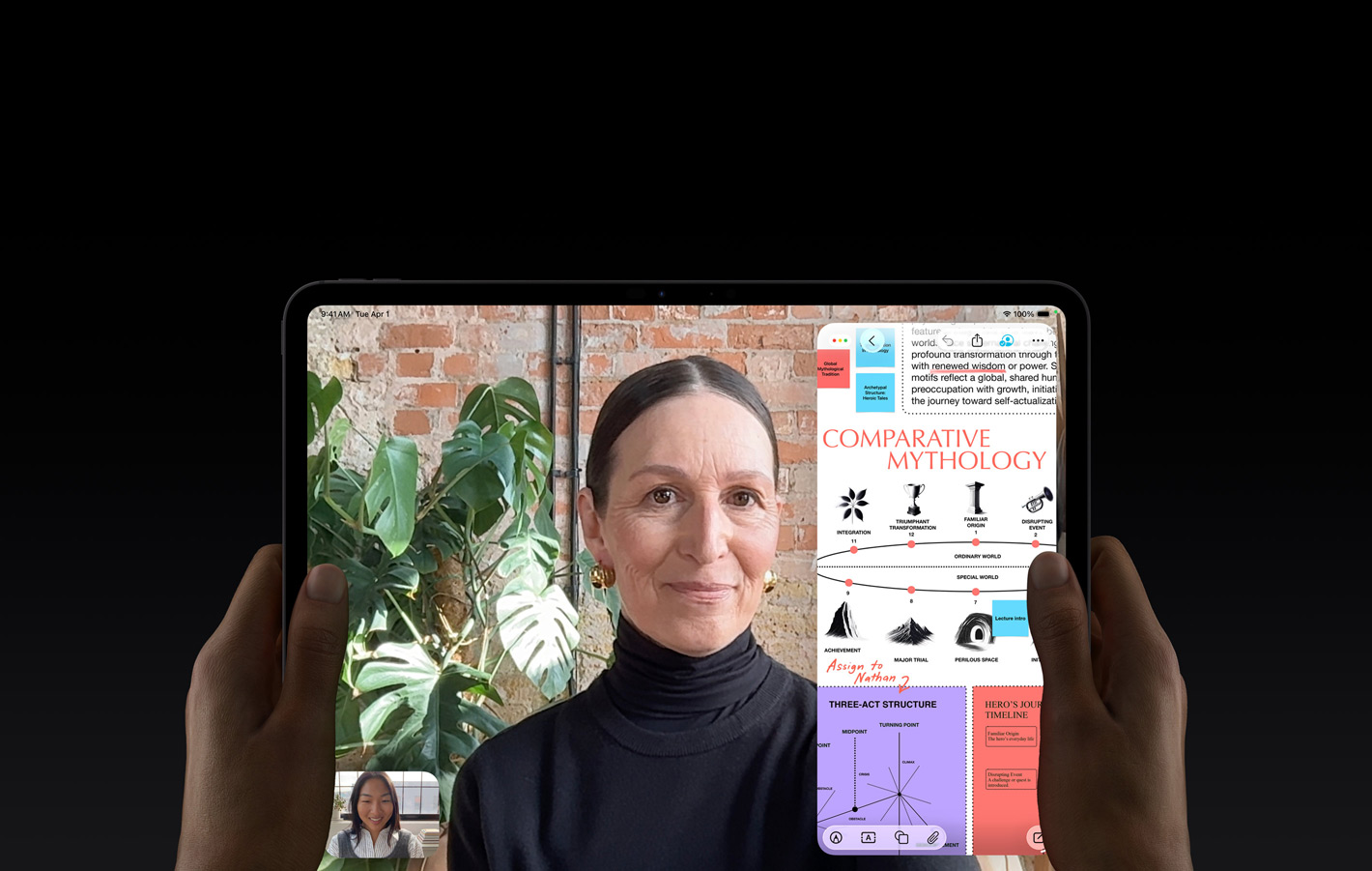 En iPad Pro i fargen stellarsvart vises forfra. To hender holder den i liggende retning. Enheten har avrundede hjørner og svart kant rundt skjermen, som viser en direkteoversatt FaceTime-samtale og en utdanningsapp. Utdanningsappen viser en presentasjon om «Comparative Mythology» med tekst, diagrammer og håndskrevne notater.