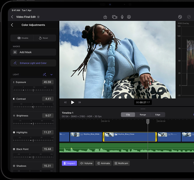 iPad Pro, front exterior, black display bezel, display showing video editing app with a picture of a model in a blue coat and black and white printed pants, color adjustment panel open on the left with various video effects and sliders, timeline at the bottom with multiple video clips and audio tracks