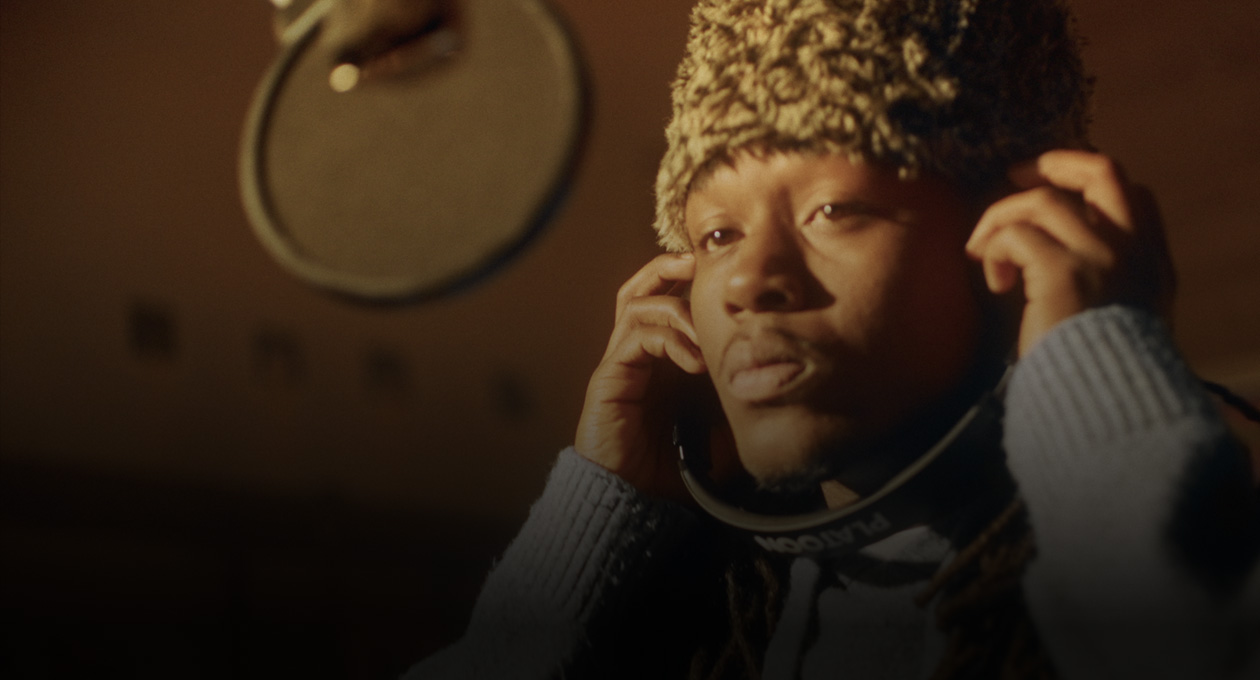 Foto Kiddo K di studio musik, berdiri di depan mikrofon rekaman dan mendengarkan melalui headphone