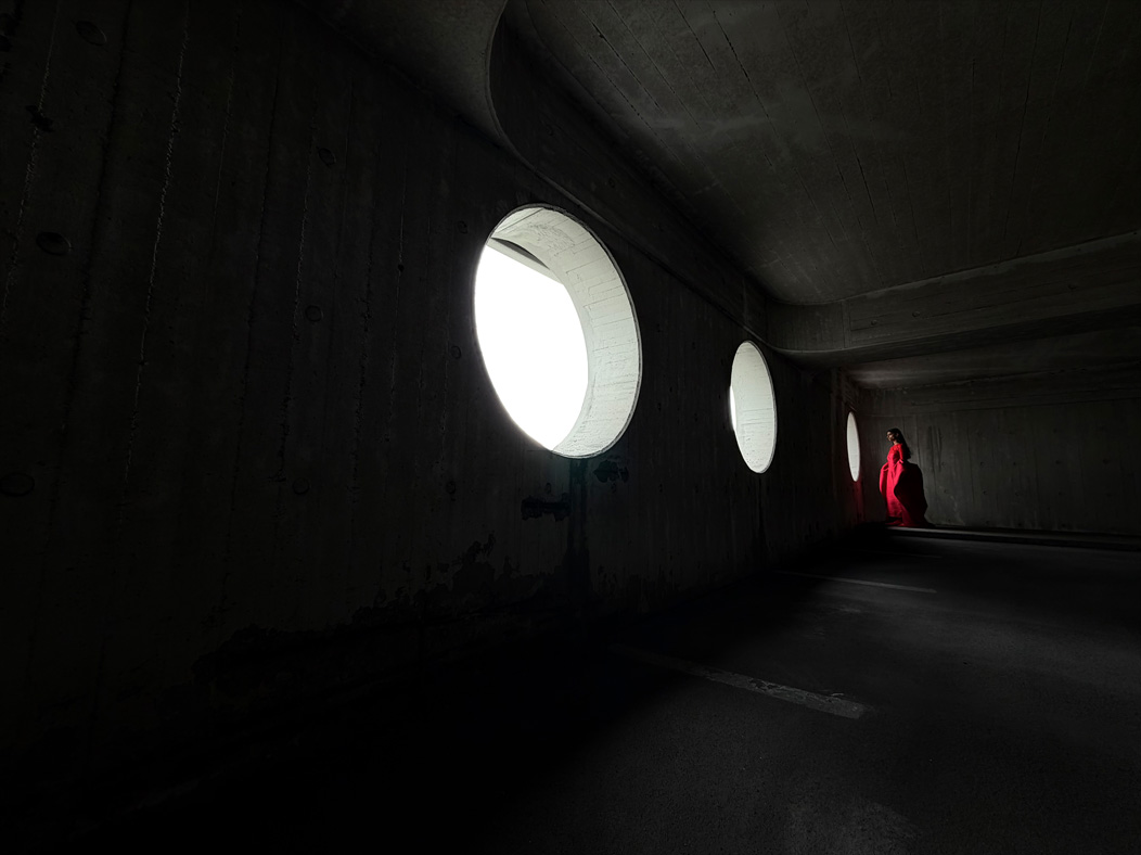 Ilgą raudoną suknelę dėvinti moteris pozuoja tuščioje patalpoje. Programoje „Nuotraukos“ perjungiami toliau pateikti židinio nuotolio nustatymai taip parodant profesionalios kamerų sistemos galimybes: 200 mm, 100 mm, 48 mm, 35 mm, 28 mm, 13 mm ir „makro“